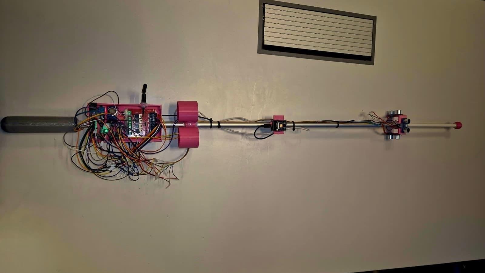 The SolCane prototype lying across a light surface, seen from above, with the handle on the left and the tip on the right. From the handle: a pink case with the control board, an antenna and loose coloured wires, two round pink housings, a small camera board halfway along the metal shaft, and the ultrasonic sensors in a pink bracket near the tip.